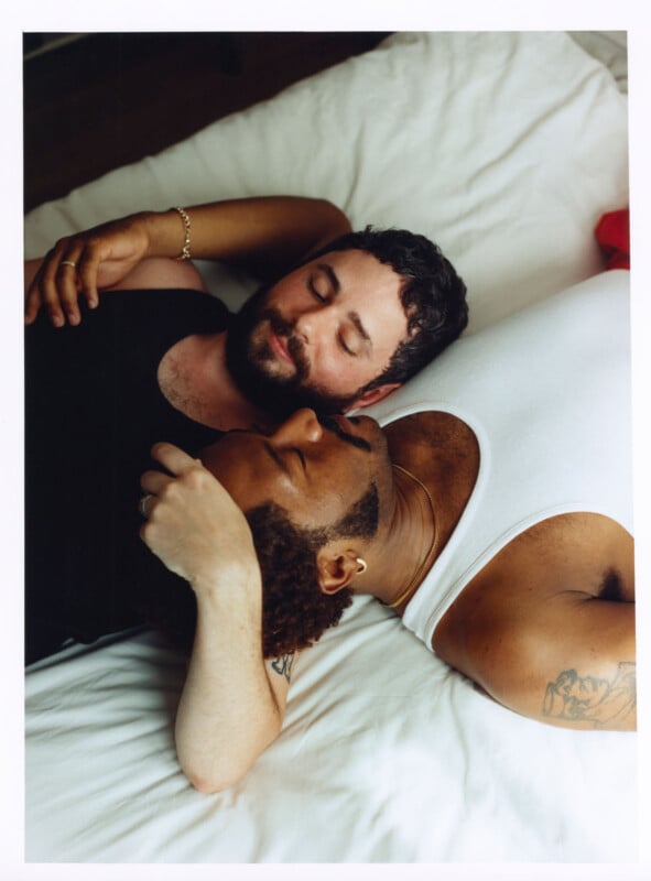Two men are lying on a bed, close together. One man with a beard and dark hair rests his arm around the other, who has short curly hair and a visible tattoo. Both appear relaxed and affectionate.