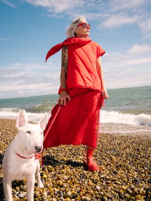 An older person with gray hair, wearing a bright red outfit and sunglasses, stands on a rocky beach next to a white bull terrier dog. The ocean and blue sky with clouds are in the background.