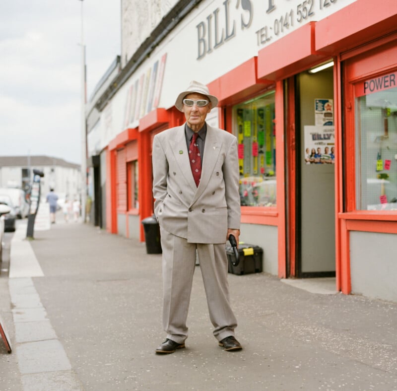 An older man in a light gray suit, tie, sunglasses, and hat stands on a city sidewalk in front of a red and white storefront called "BILL'S." He holds a black bag and looks toward the camera.