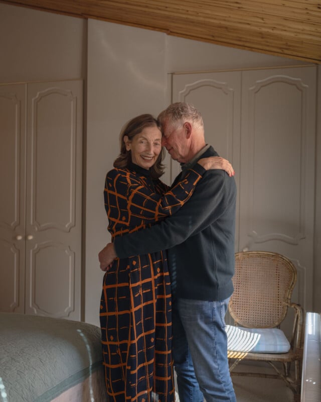 An older couple embraces warmly in a cozy, sunlit bedroom. The woman wears a checked dress and smiles, while the man gently leans his head to hers. A wicker chair and bed are visible in the softly lit room.