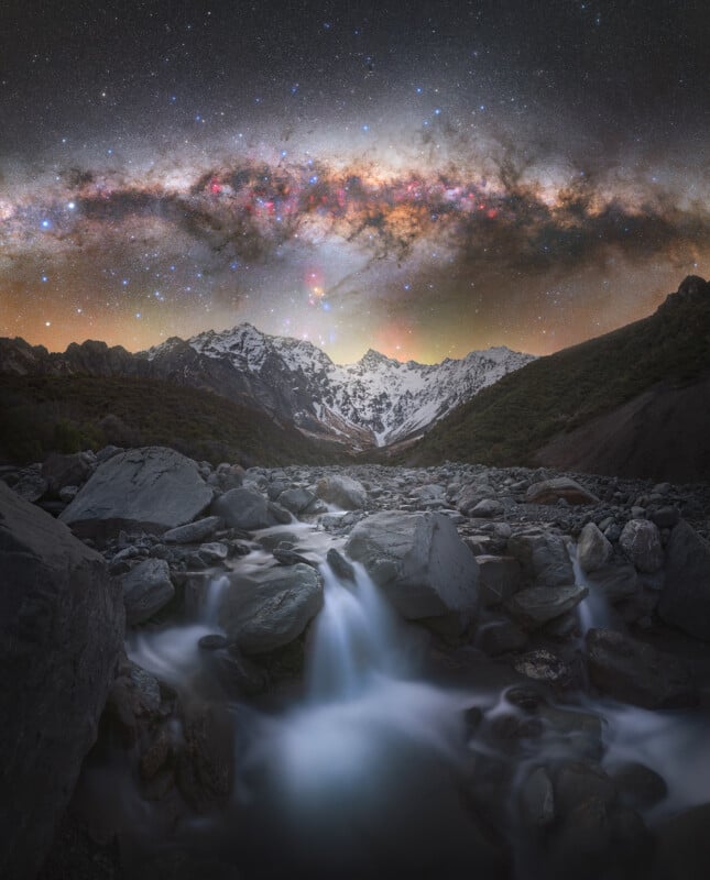 A rocky mountain stream flows in the foreground, framed by snow-capped peaks. Above, the vibrant Milky Way stretches across a clear night sky filled with stars.