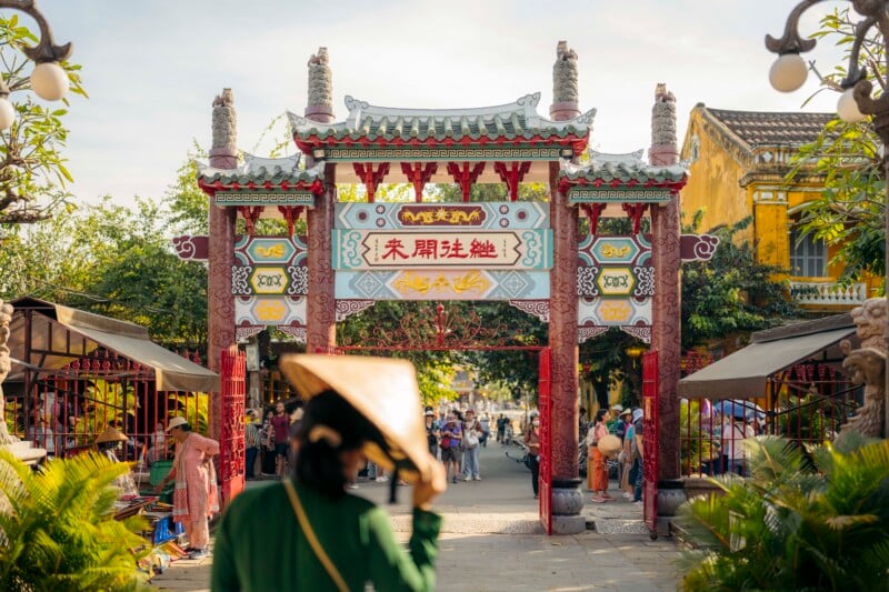 A person in a traditional conical hat walks toward an ornate, colorful Asian gate with decorative patterns and Chinese characters. The scene is lively, with people and market stalls on both sides. Sunlight brightens the setting.
