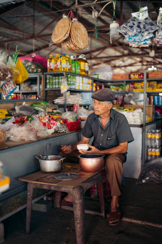 An elderly man in a cap sits at a small table eating food from a bowl in a bustling market stall filled with various groceries, snacks, and household items. Pots and utensils are on the table in front of him.