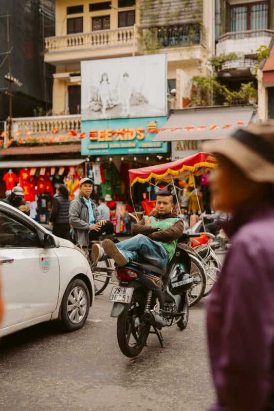 A man relaxes on a parked motorbike in a busy city street, with cars, people, and colorful storefronts in the background. Another person is seen walking by in the foreground, slightly out of focus.