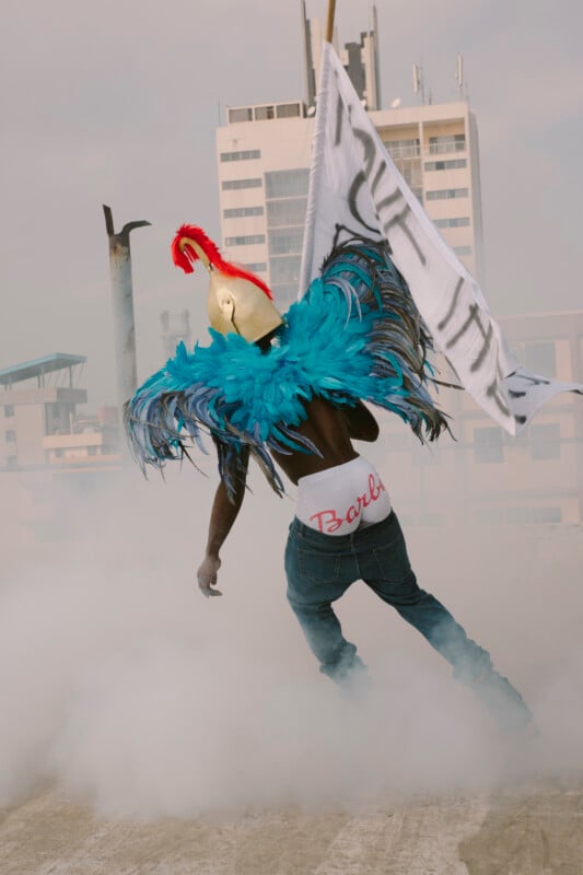 A person wearing a gold helmet, blue feathered wings, and "Barbie" underwear stands in jeans on a rooftop surrounded by smoke, holding a large white flag with buildings in the background.