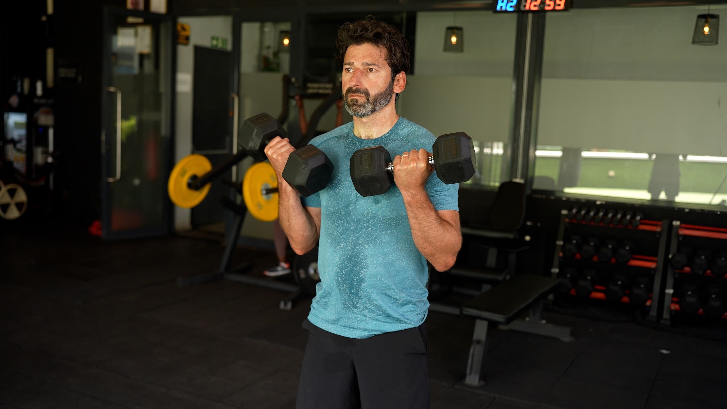 as part of a longevity workout plan, a man with dark hair wearing a blue shirt and black shorts demonstrates how to do biceps curls with a dumbbell