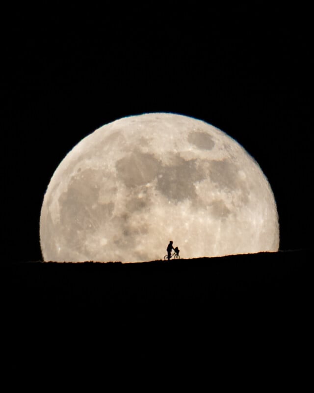 A large, bright full moon dominates the night sky as a silhouette of a person riding a bicycle appears against its lower edge on a dark horizon.