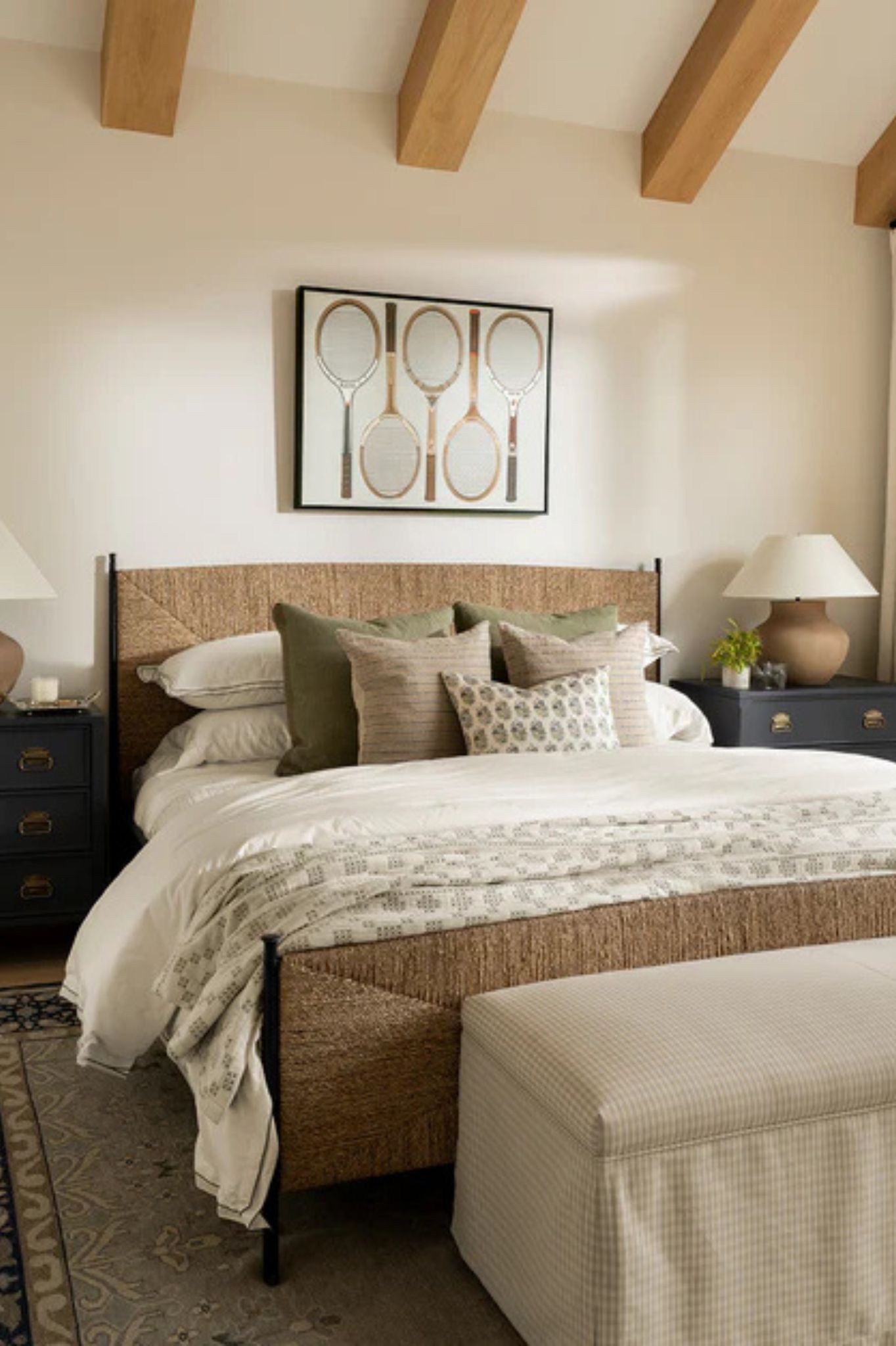 Bedroom with beamed ceiling, rattan bed frame with linen bedding and print of tennis rackets above bed