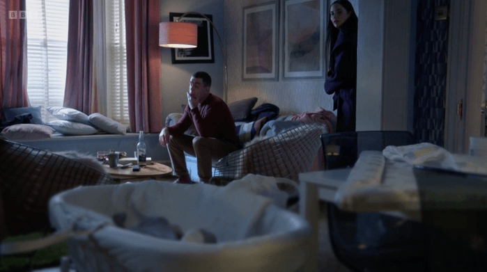 A baby's cot in the foreground with Scott Maslen as Jack Branning sat in his living room in the background with Jacqueline Jossa as Lauren Beale standing in EastEnders.