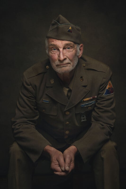 An elderly man wearing a vintage military uniform and cap sits facing forward with hands clasped, gazing intently. Dark, neutral background highlights his thoughtful expression and decorated jacket.