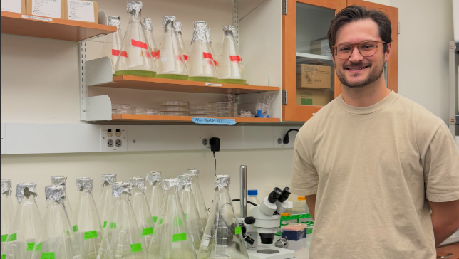 Evan Stair in a laboratory beside several flasks containing green liquid