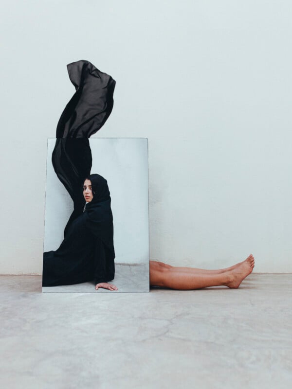 A mirror on the floor reflects a woman draped in flowing black fabric, with part of the fabric billowing upward; her bare legs extend from behind the mirror on a concrete surface against a plain wall.