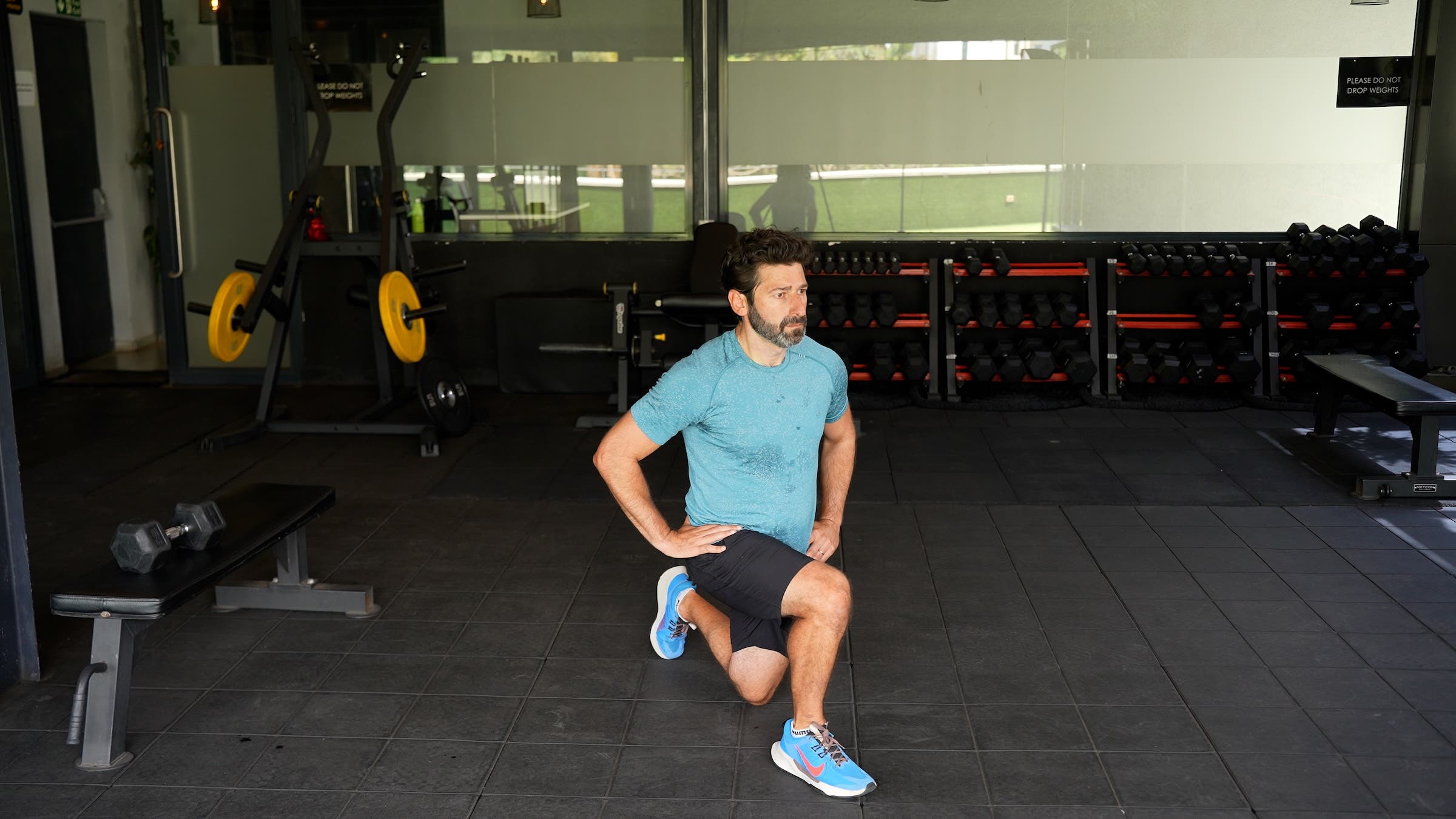 a man with dark hair wearing a blue shirt and black shorts does a lunge in a gym