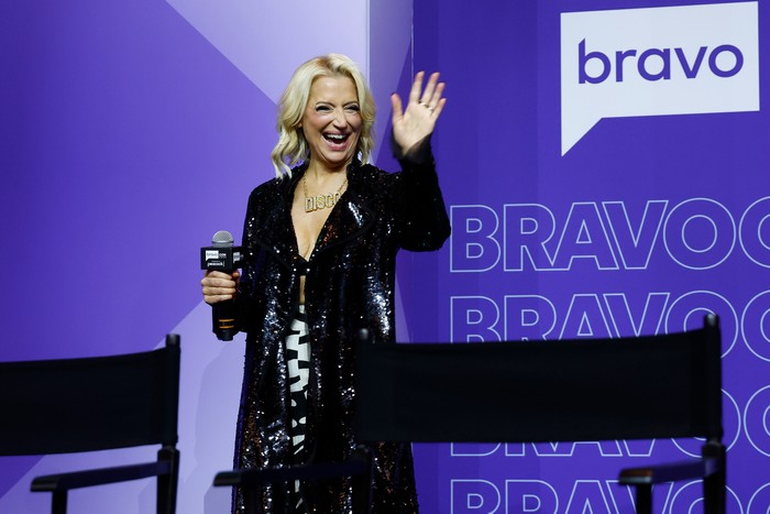 Dorinda Medley smiling ahead, holding a microphone with one hand and waving with the other.