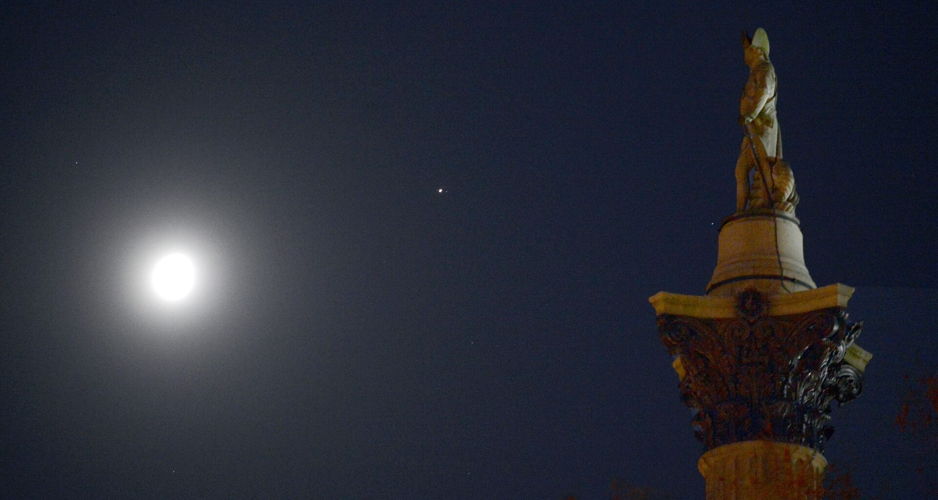 The moon is pictured shining in the night sky with a bright point of light representing Jupiter to its upper left. A stone statue of a military figure atop an ornate column is positioned to the right of the shot, appearing to face the solar system pair.