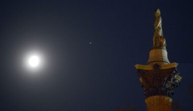The moon is pictured shining in the night sky with a bright point of light representing Jupiter to its upper left. A stone statue of a military figure atop an ornate column is positioned to the right of the shot, appearing to face the solar system pair.
