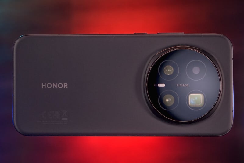 A black Honor smartphone with a large circular camera module is shown against a blurred red and dark background. The phone is positioned horizontally with the camera module on the right.