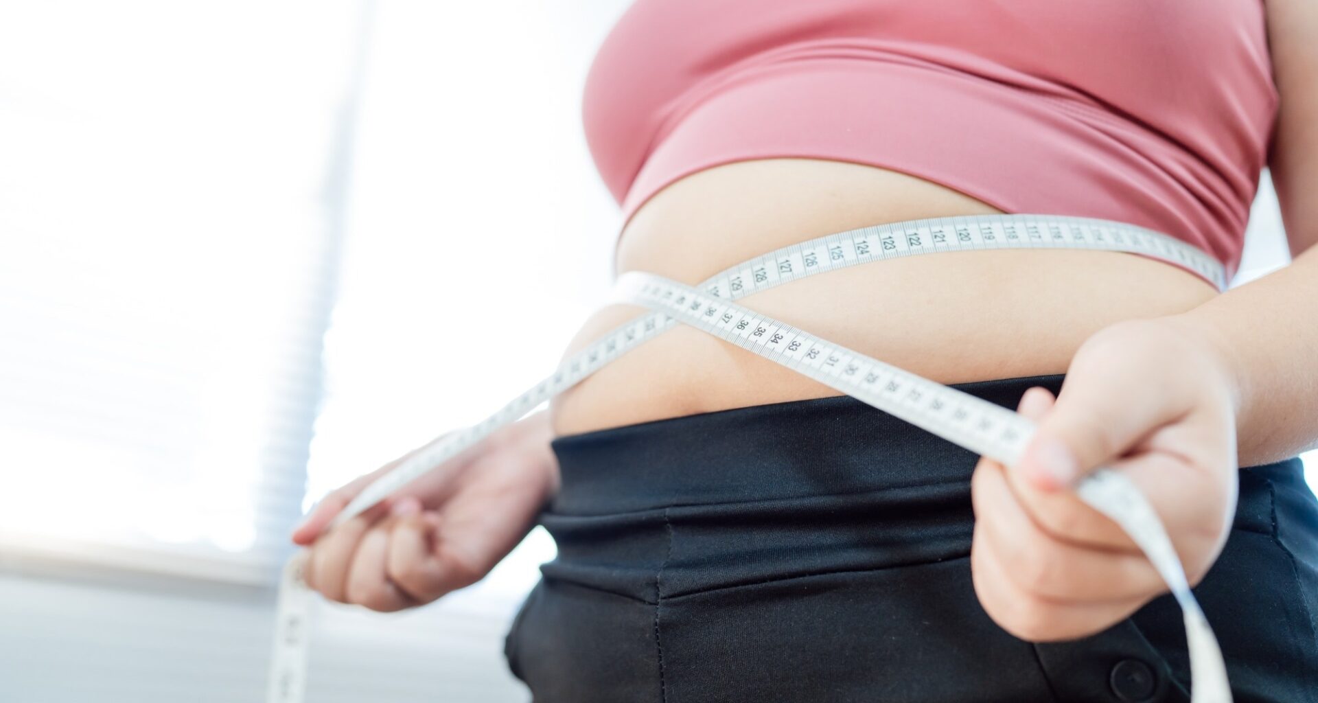Overweight woman in pink sports top pulling measuring tape around waist