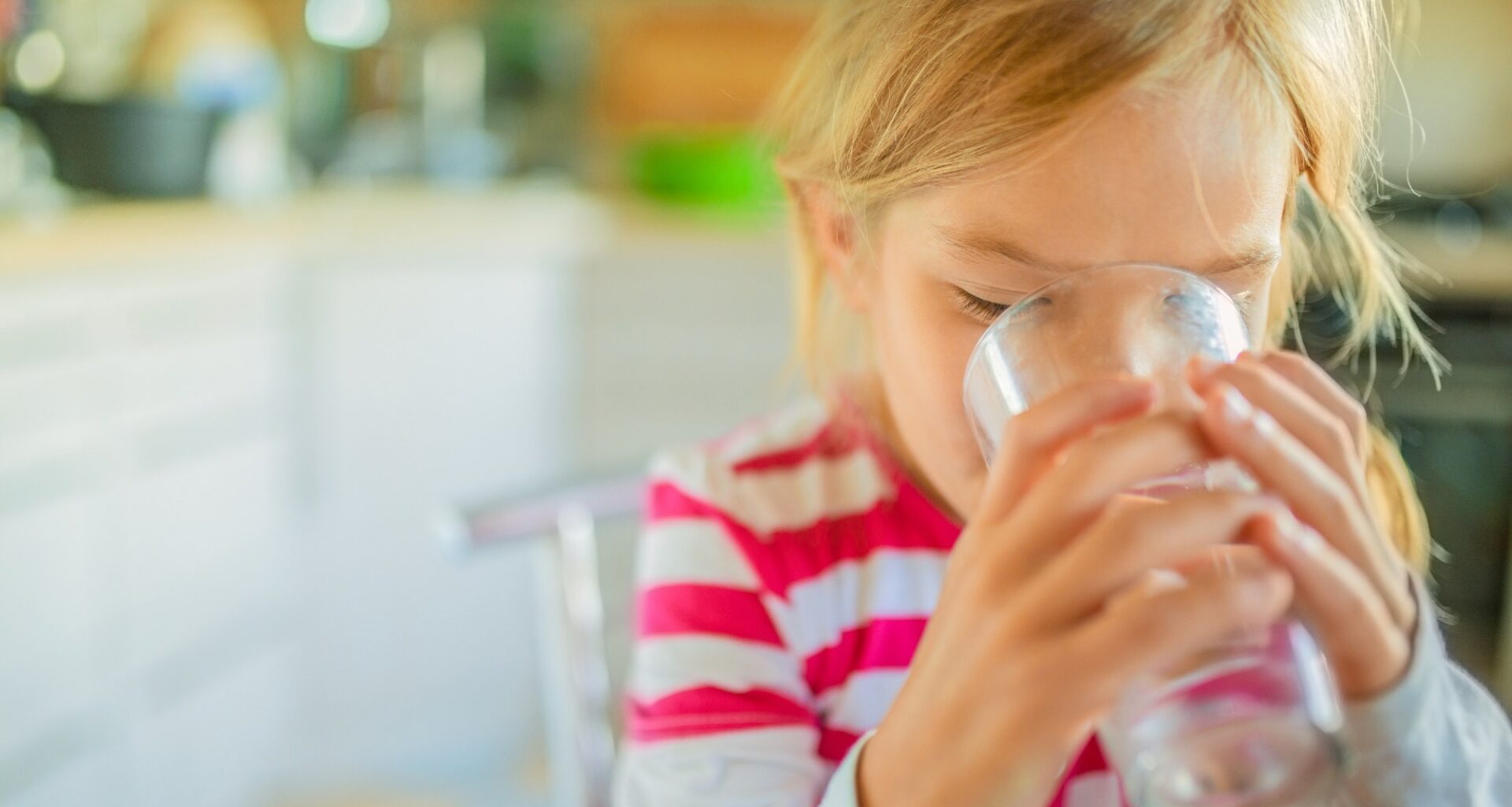 Beautiful smiling little girl drinking a glass of water against the background of the kitchen.