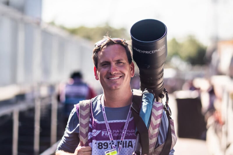 A smiling man wearing a press badge carries a large camera with a telephoto lens over his shoulder, standing outdoors in a sunlit, busy environment.
