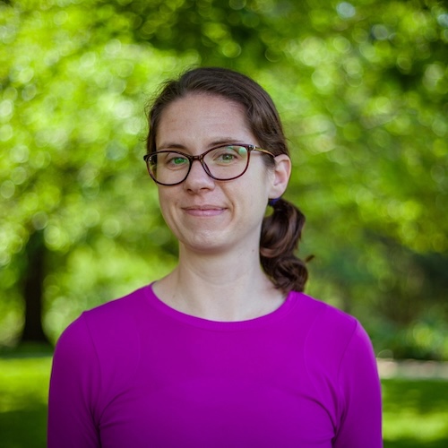 Smiling woman with eyeglasses and dark hair tied behind her head, wearing a purple shirt.