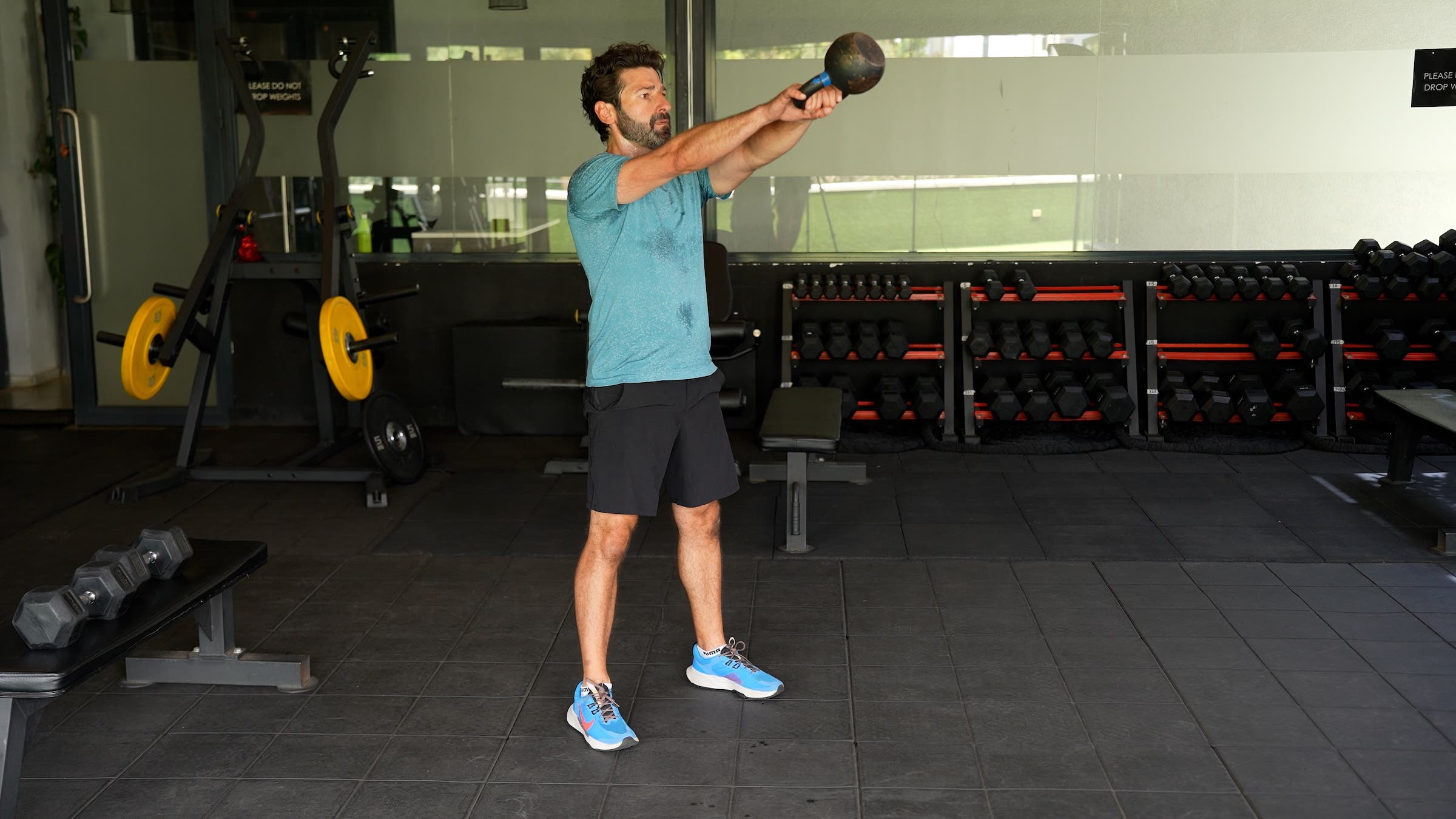 a man with dark hair wearing a blue shirt and black shorts demonstrates a kettlebell swing 