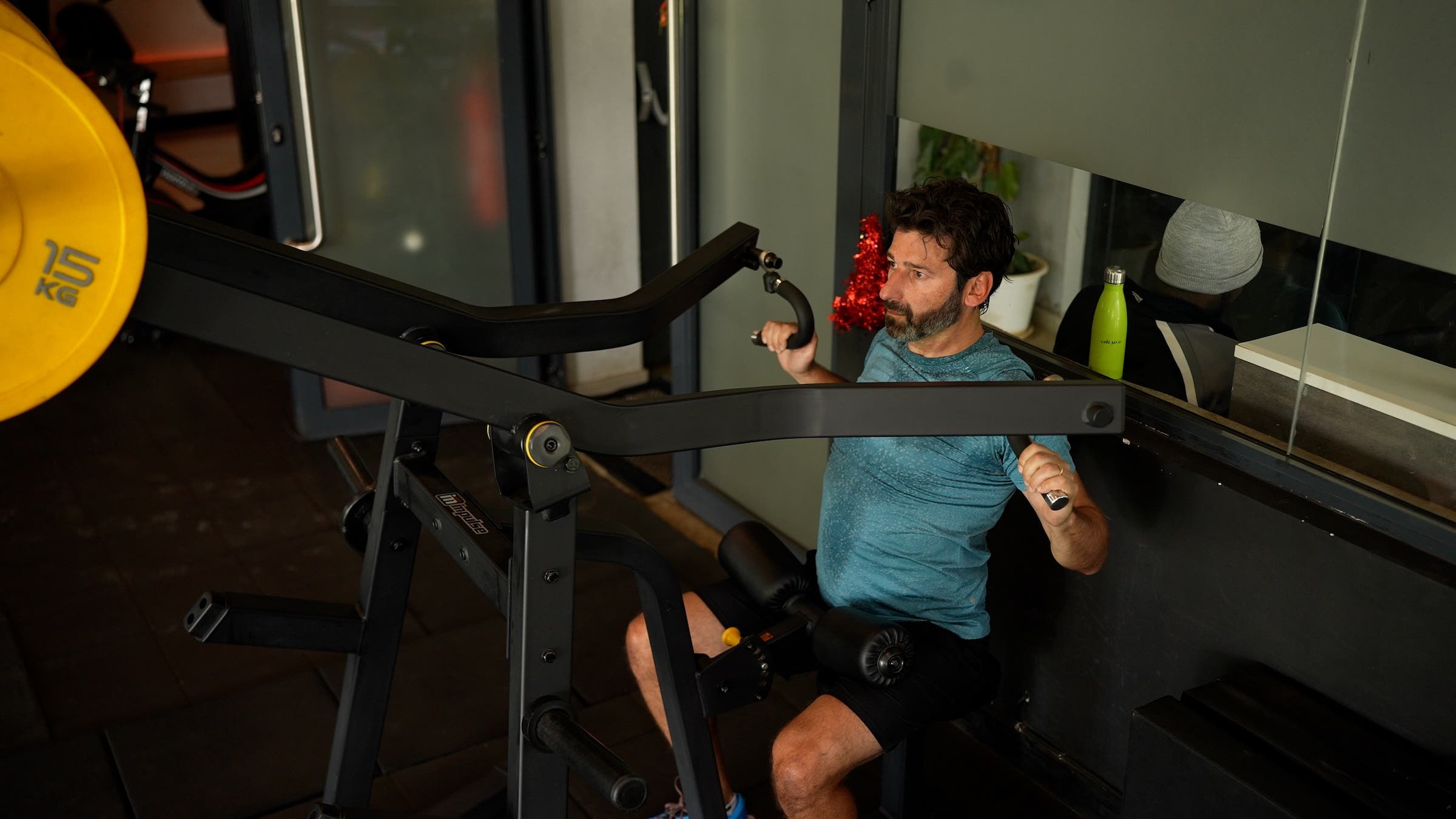 a man with dark hair wearing a blue shirt and black shorts does a lat pull-down as part of a longevity workout plan