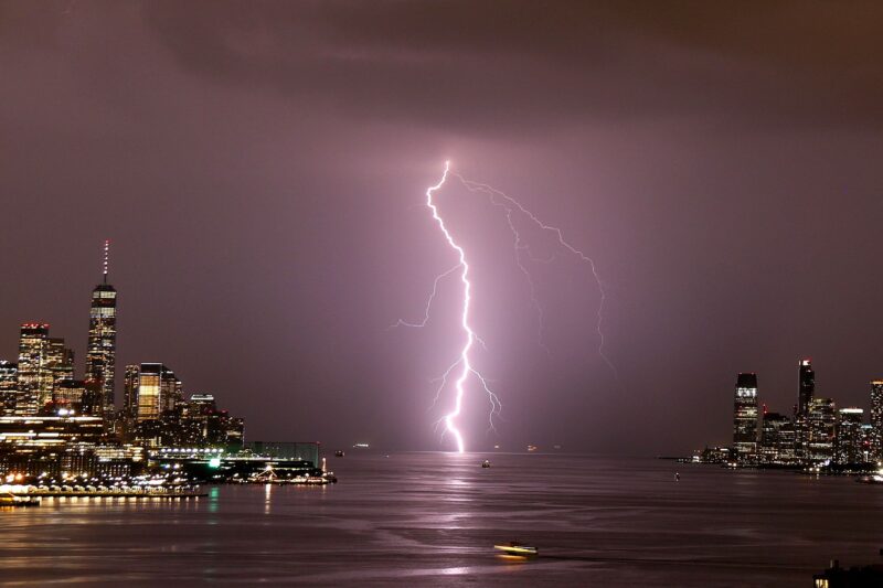 Lightning forms: A jagged, glowing, vertical white line striking the water between two areas of tall buildings.