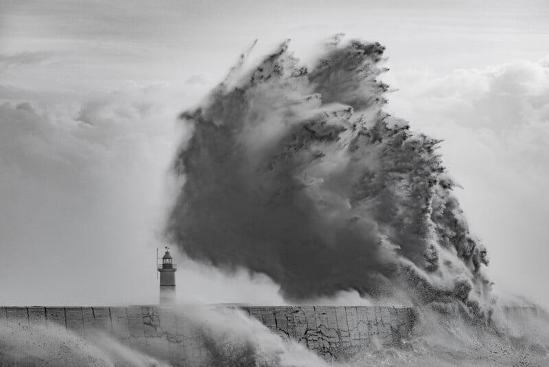 A massive wave crashes dramatically against a breakwater, dwarfing the small lighthouse standing on the pier, with water spraying high into the cloudy sky. The image is black and white.