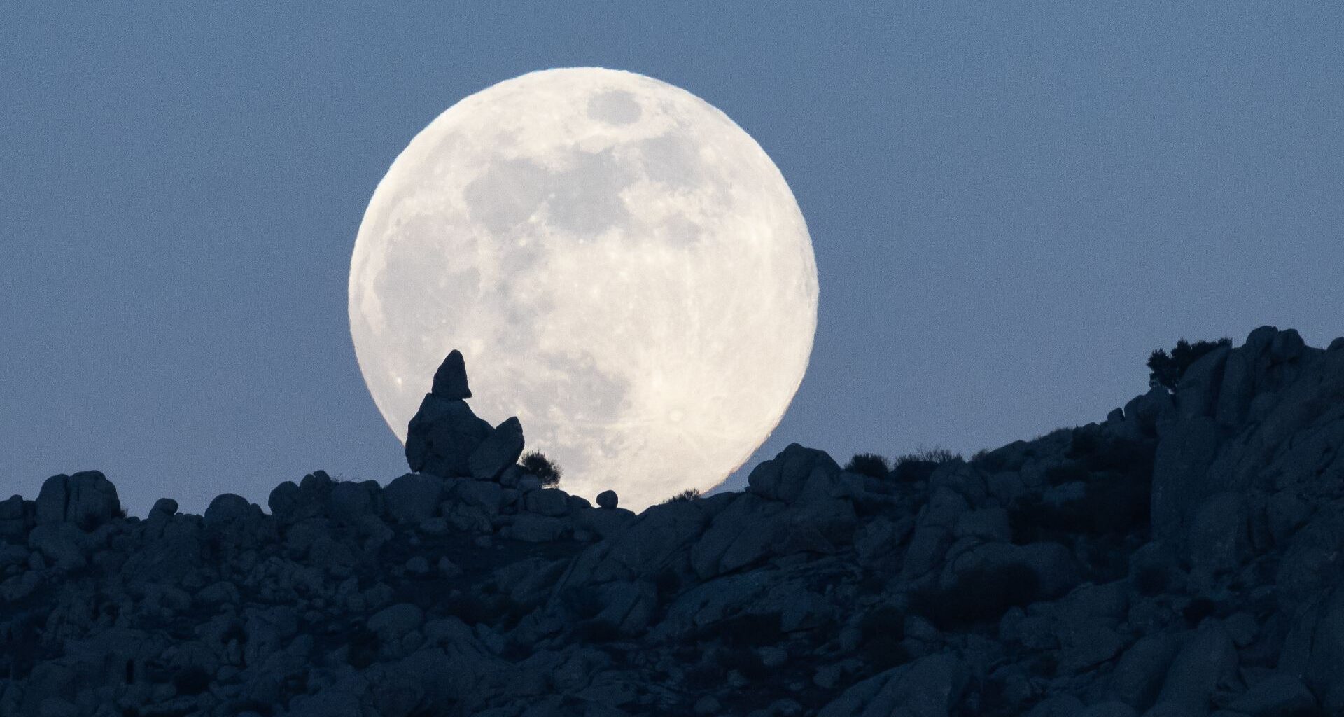 A full moon is photographed rising above a rocky hillside in the dark blue evening sky.