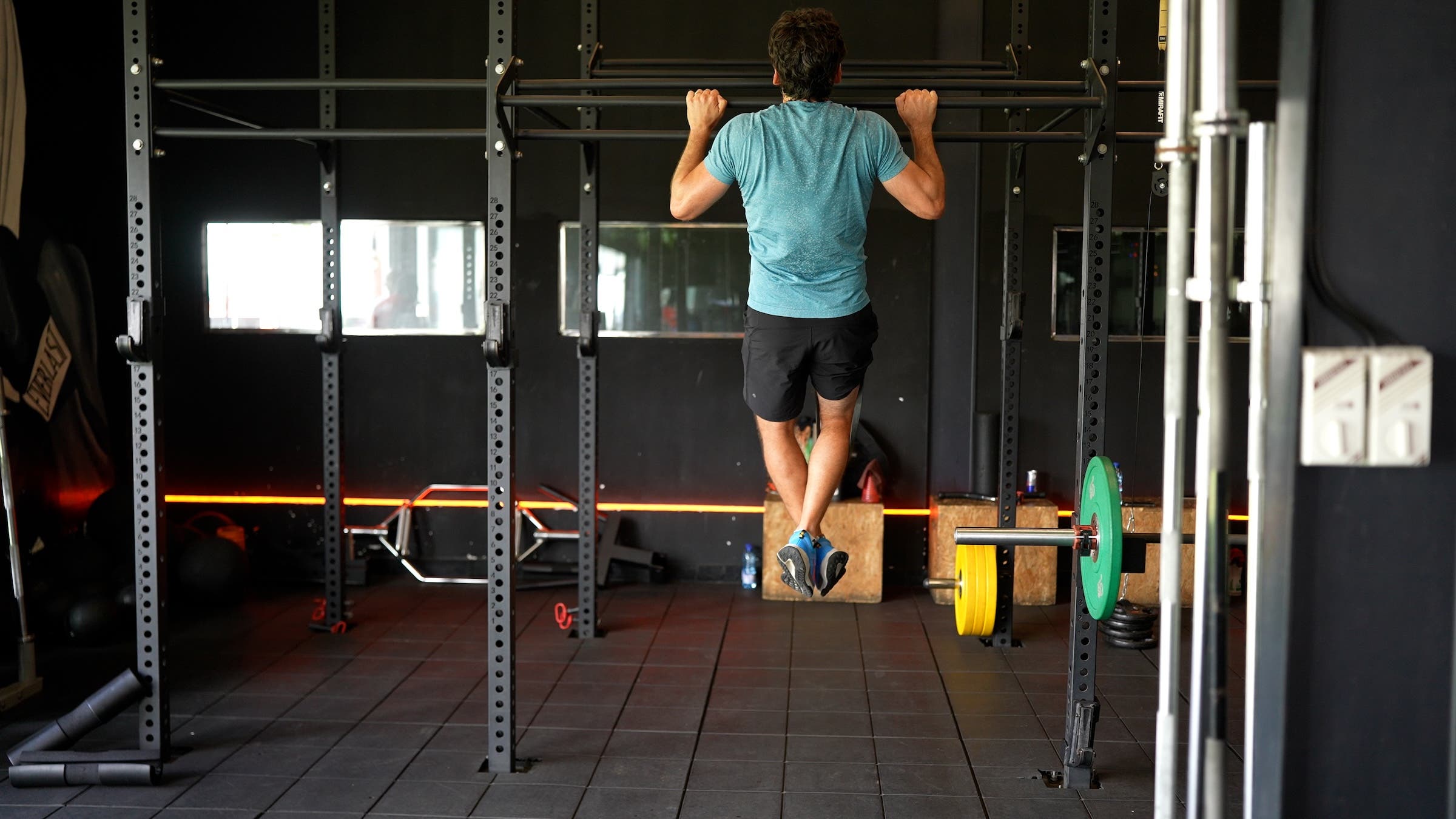from behind, a man with dark hair wearing a blue shirt and black shorts does a pull-up on a pull-up bar