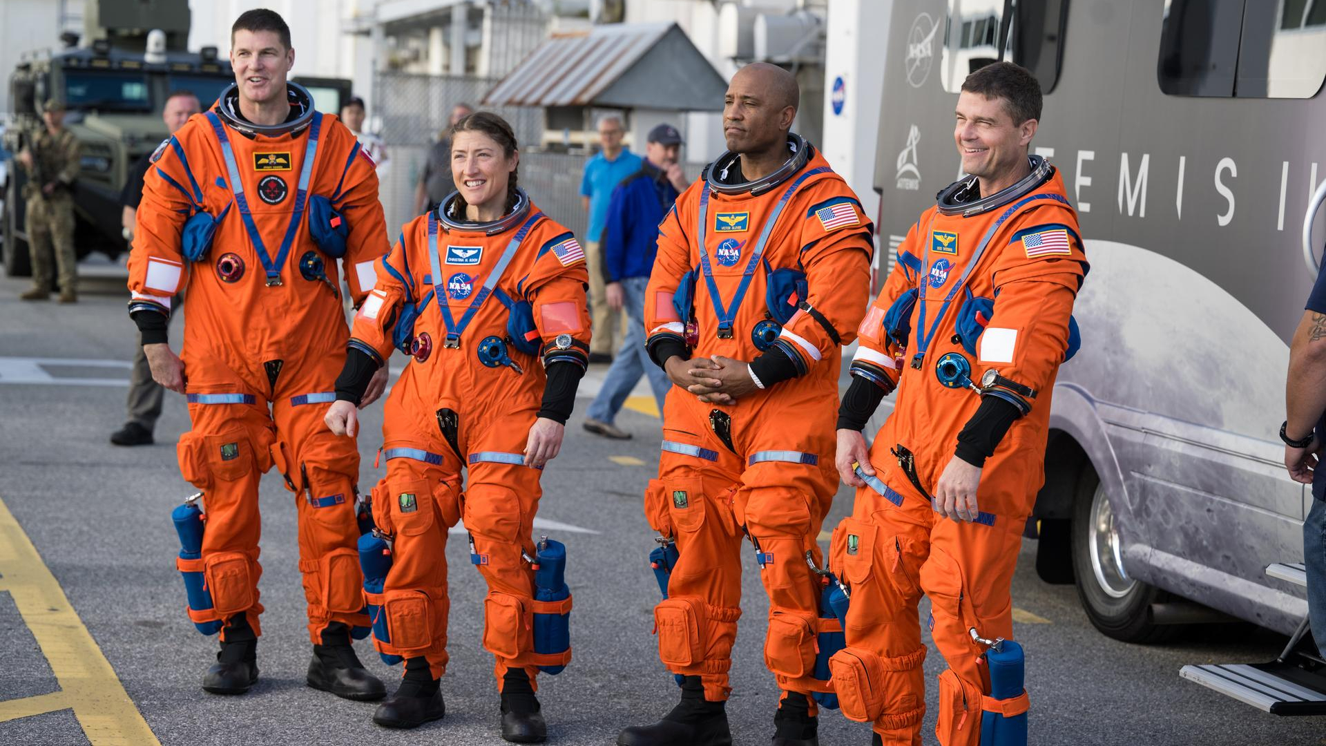 Four people in orange space suits stand on a road in front of a large van and look at a crowd off camera