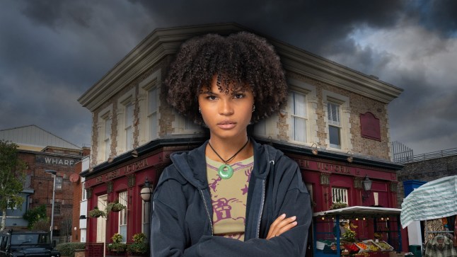 EastEnders' Jasmine Fisher standing in front of the Queen Vic pub with her arms folded. The sky above is dark and cloudy.