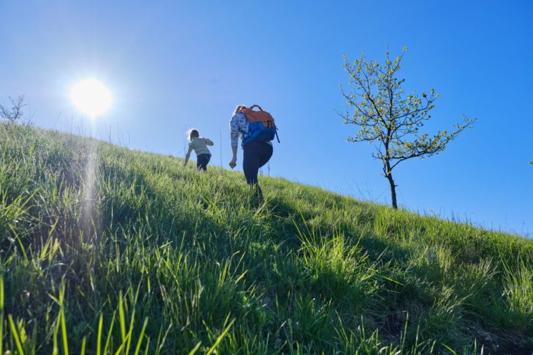 A mother and child hike up a grassy hill bathed in bright sunlight, surrounded by vibrant green grass and a clear blue sky. The dynamic low-angle view conveys energy, determination, and the joy of movement in nature.