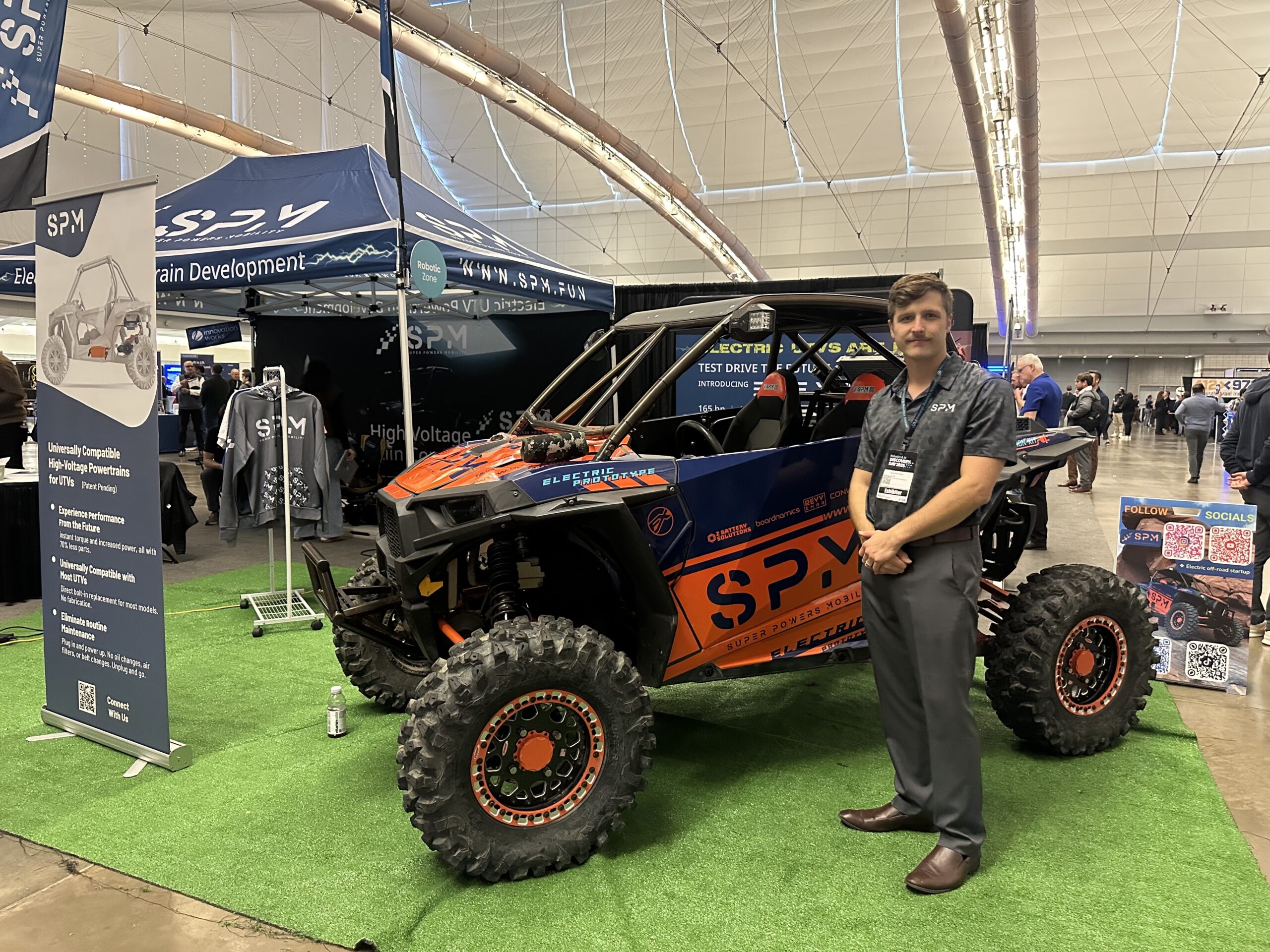 A man stands next to an off-road vehicle on display at a trade show booth, featuring banners and tents with SPV branding.