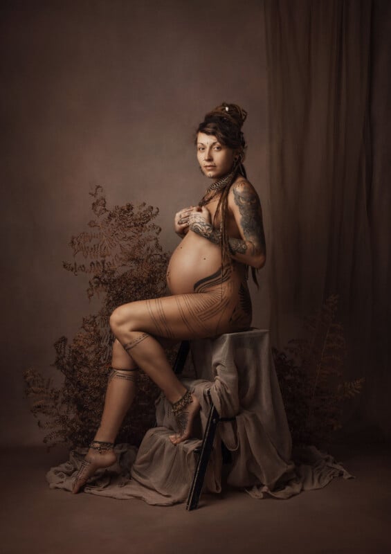 A pregnant person with long hair and tattoos poses seated on a draped stool, one hand on their belly. They wear sheer clothing and are surrounded by dried ferns against a muted brown backdrop.
