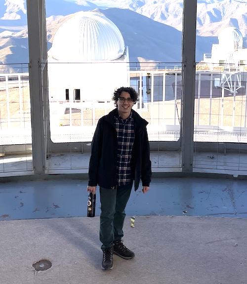 Man with curly hair, checkered shirt and open jacket standing in a room with huge glass windows. An observatory dome and background mountains are seen outside.