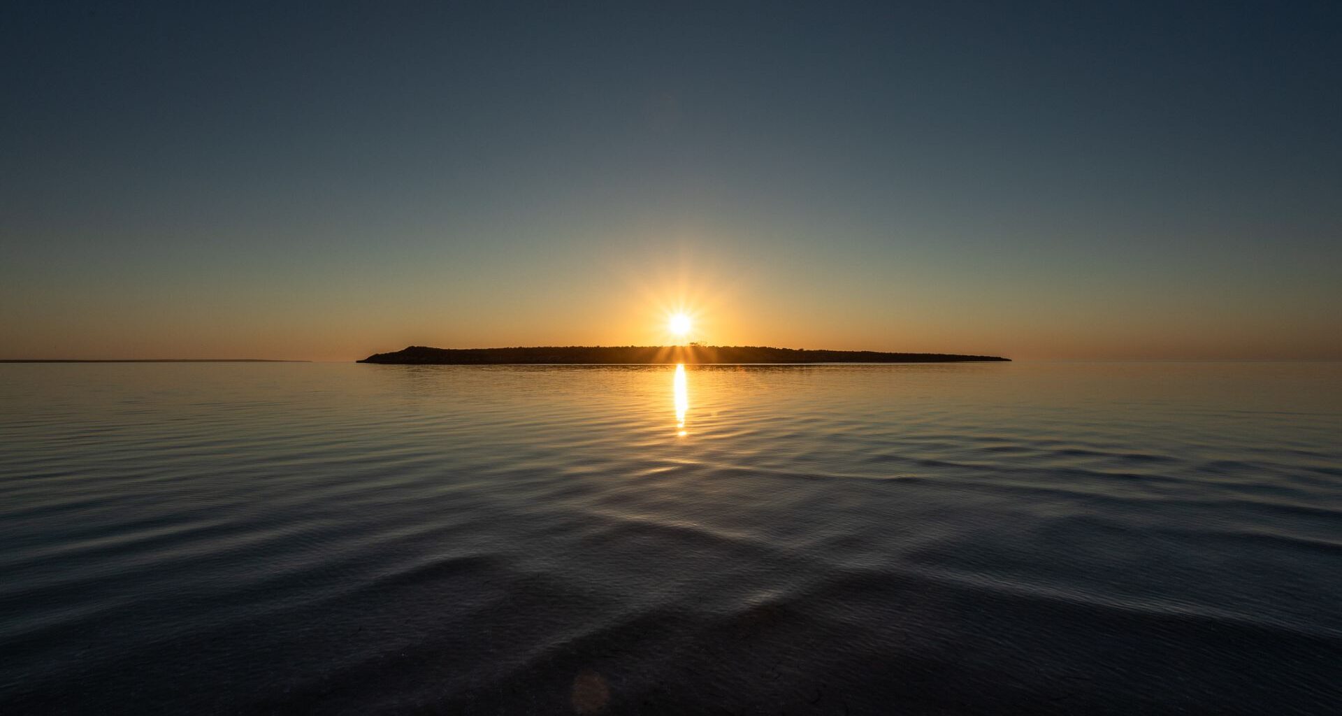 A sunset over a lake with a silhouette of an island right underneath the setting sun