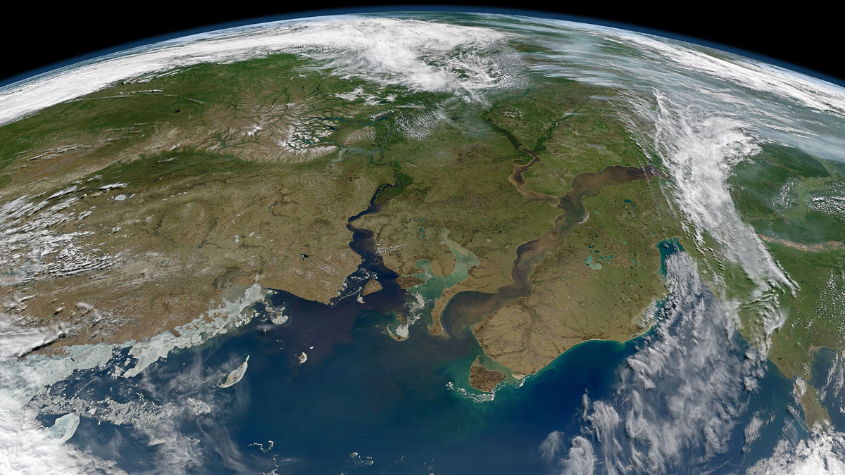 A satellite view of a portion of Earth shows the planet’s curved horizon at the top of the image. Green and brown landmasses are cut by two large, blue, winding rivers that empty into the ocean in the foreground. Clouds are visible on the edges of the image.
