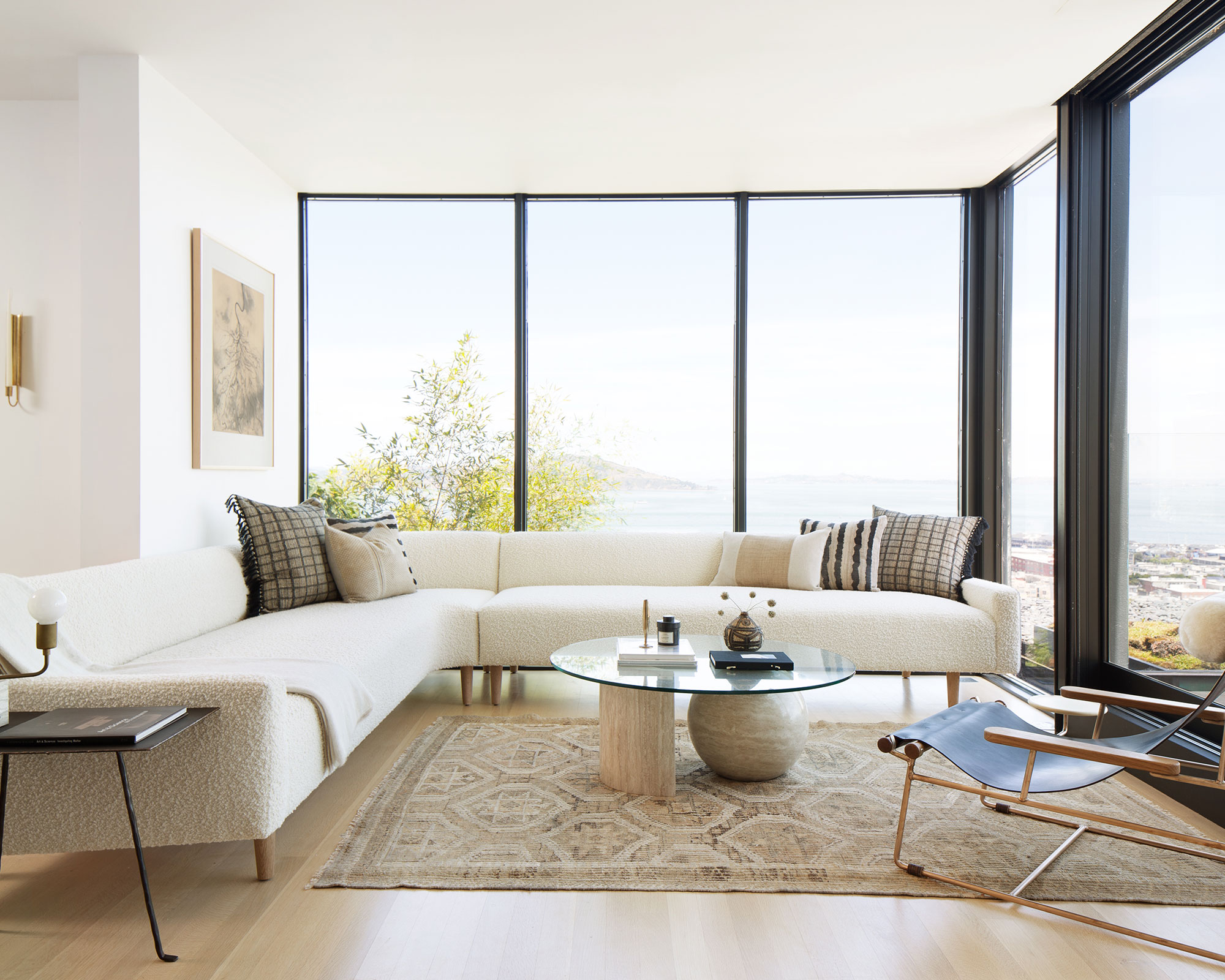 A sun-drenched, modern living room with floor-to-ceiling windows overlooking a coastline. A large, cream-colored boucl&eacute; sectional sofa spans the width of the room, adorned with textured plaid and striped pillows. In the center, a round glass coffee table sits on a travertine and stone pedestal base over a vintage-style patterned rug. To the right, a black leather and wood sling chair faces the view, and a thin black side table stands to the left. The space is bright, airy, and minimalist in design.