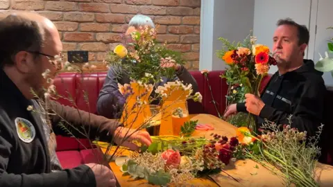 Three men sit round a pub table holding flowers that they are arranging in their hands. On the table are more flowers and pints of beer.