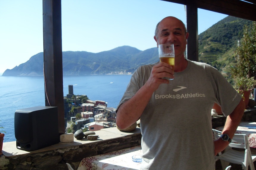 A man standing with a beer in front of a view of a bay surrounded by mountains covered in trees.