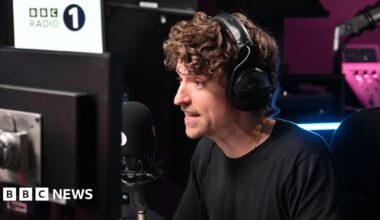 Greg James speaking into a black radio microphone with large black headphones over his head. He is talking and staring ahead which is to the left of the camera. There is a computer monitor in front of him to the right and a large BBC Radio 1 white sign to the left. There is a pink light behind him illuminating a sound board, which is slightly out of focus.