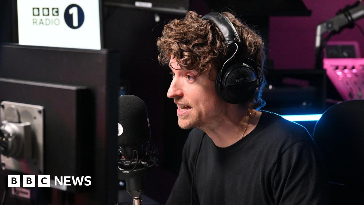 Greg James speaking into a black radio microphone with large black headphones over his head. He is talking and staring ahead which is to the left of the camera. There is a computer monitor in front of him to the right and a large BBC Radio 1 white sign to the left. There is a pink light behind him illuminating a sound board, which is slightly out of focus.
