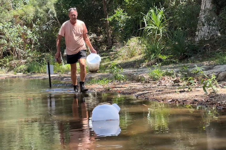 A man wades through ankle-deep water, holding a plastic trap full of tadpoles and water