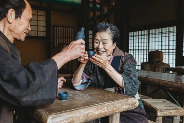 Two people in traditional attire share a joyful moment while toasting with sake cups in a wooden, Japanese-style room