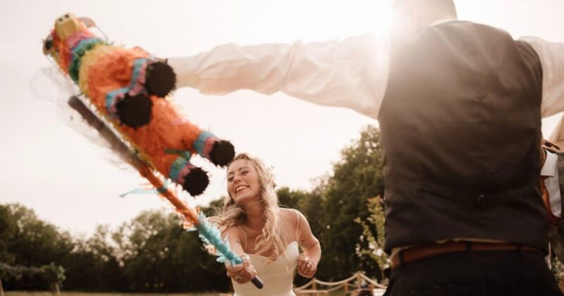 A smiling bride swings a bat at a colorful piñata held by someone in a vest and dress shirt during an outdoor celebration, with sunlight streaming and trees in the background.