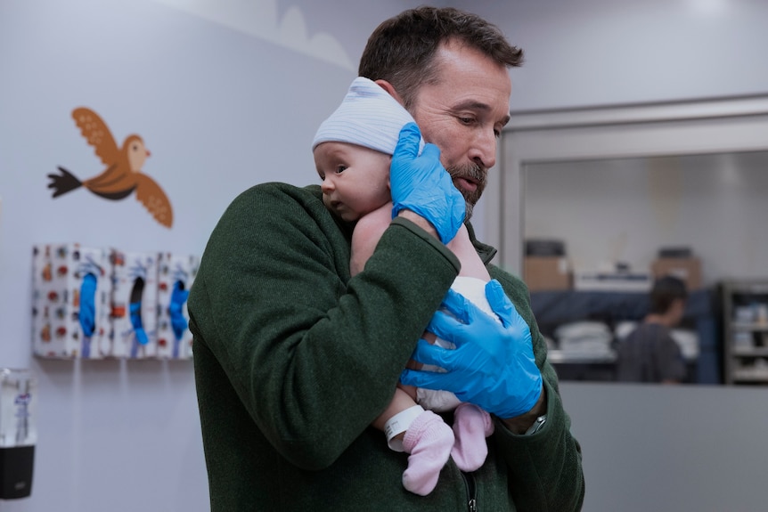 A TV still of Noah Wyle, 54, in surgical gloves, holding a baby to his shoulder in a kids room in an emergency department.