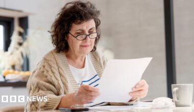 An older woman wearing glasses sits at a table looking at a piece of paper.