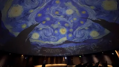 BBC A projection of Vincent van Gogh’s “Starry Night” fills a curved planetarium dome, showing swirling blue skies, bright yellow stars and the dark silhouette of cypress trees in the foreground.